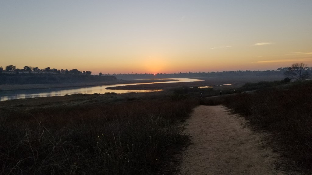 A path leads through a field towards a body of water with the sun setting in the distance.