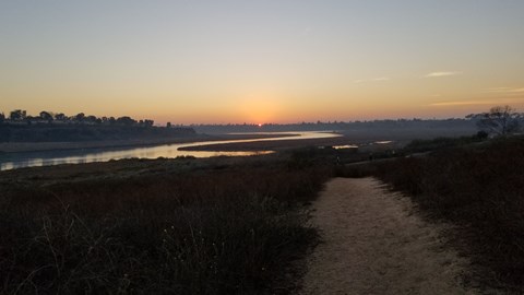 A path leads through a field towards a body of water with the sun setting in the distance.