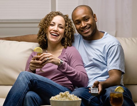 A couple is sitting on a couch, eating popcorn and drinking beverages while watching television.