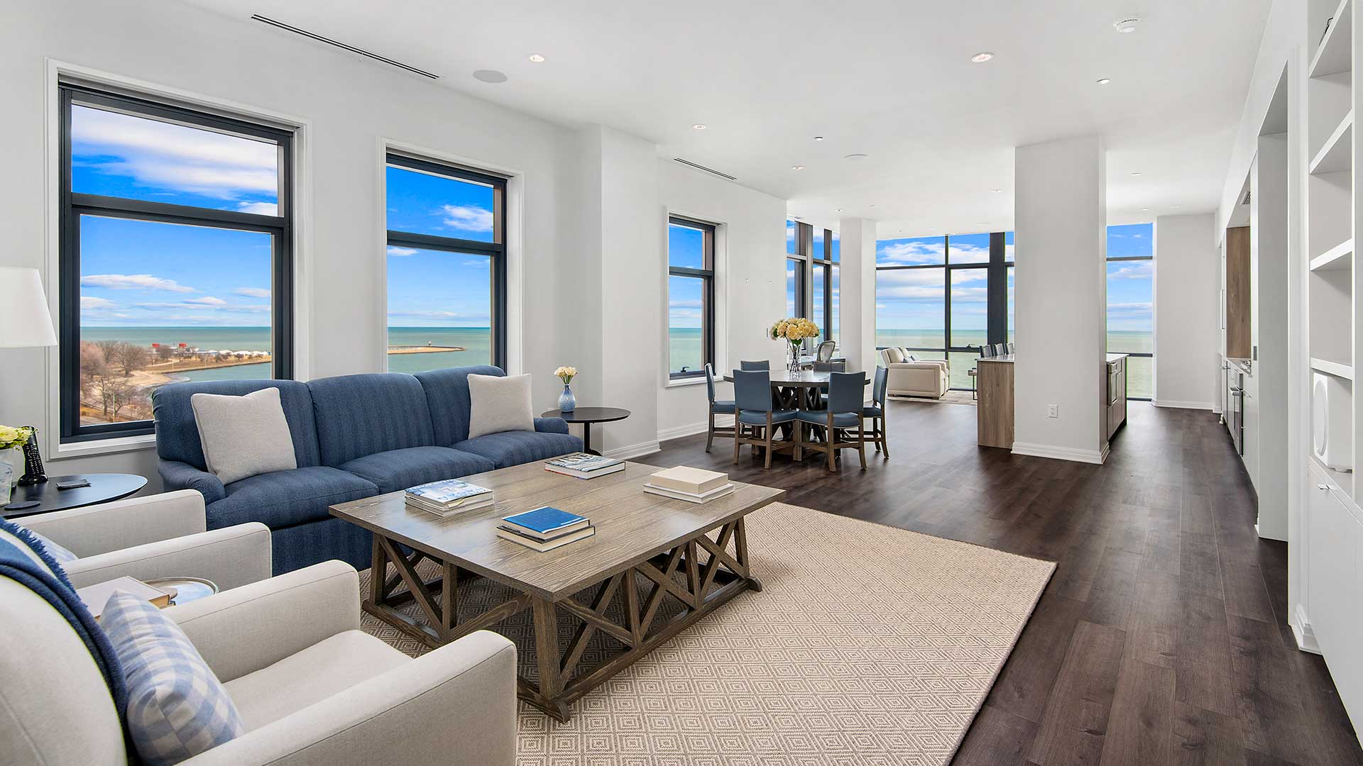 Looking through the open living room and kitchen of an open floor plan. The living room set is to the left with the dining set further down. The kitchen is down on the right. Windows line the walls an