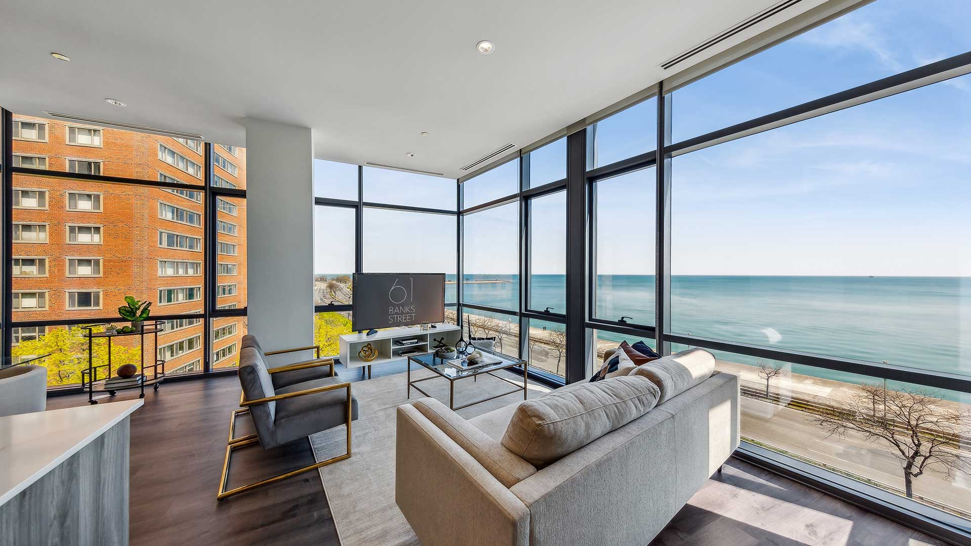 A modern living room with large windows offers a view of a waterfront and street. The room is furnished with a gray couch, chairs, and a television. An adjacent building is visible through the windows