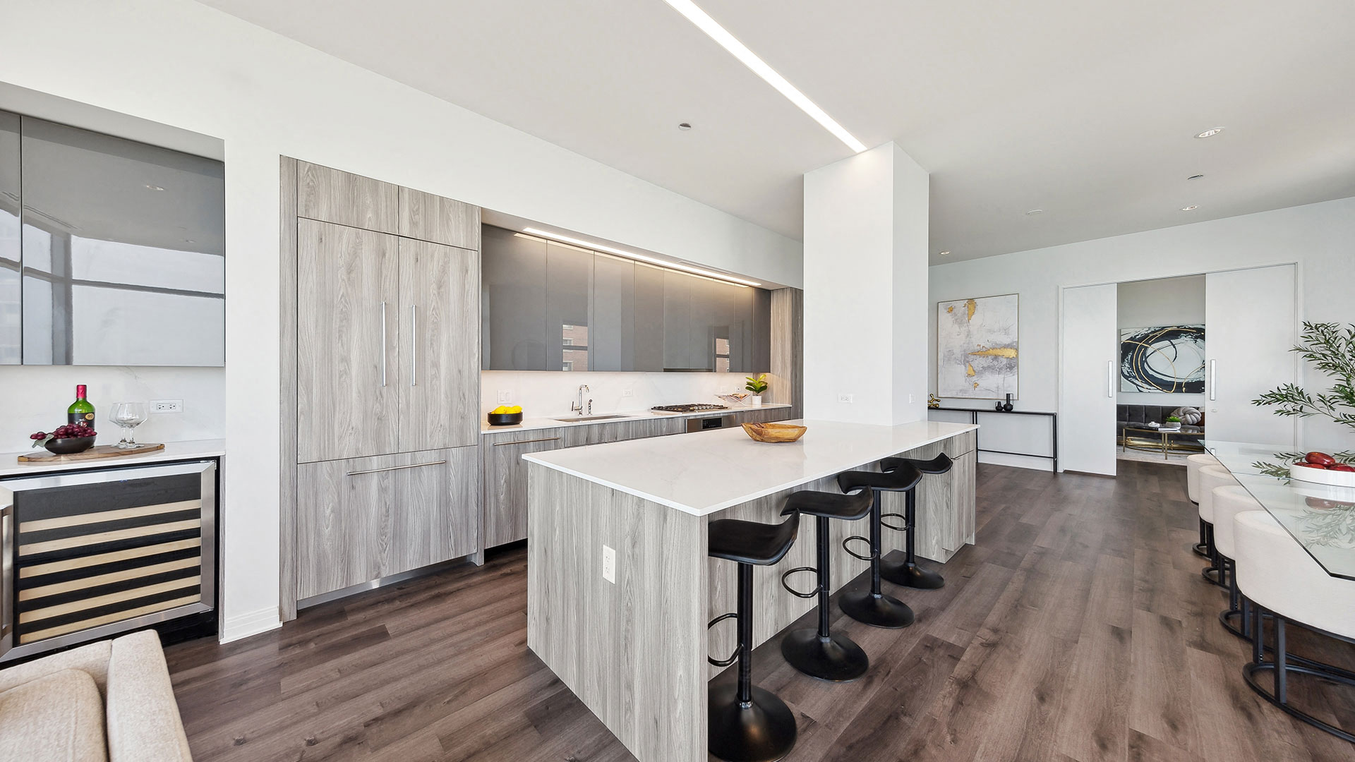 Modern kitchen with light wood cabinetry, large island with white countertops, barstools, built-in fridge, wine cooler, and contemporary decor.