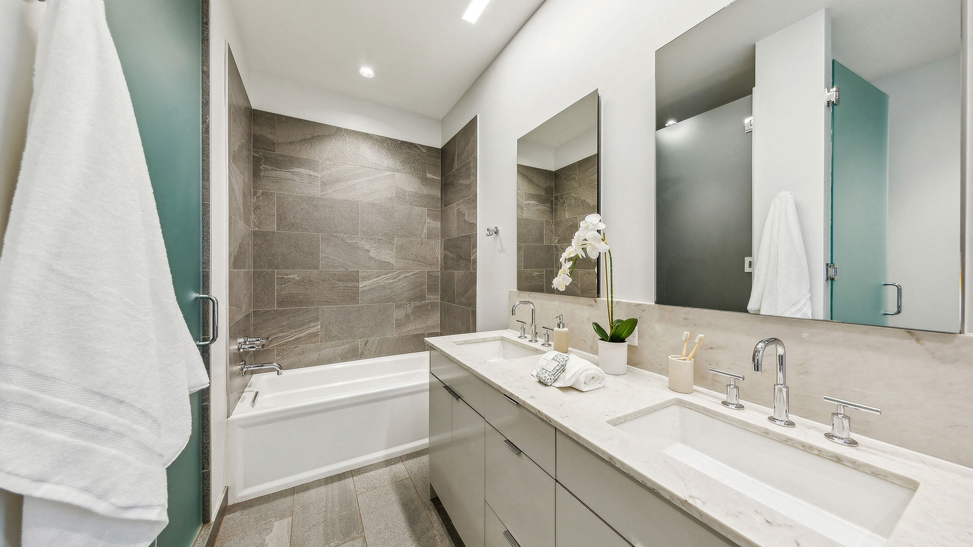 Modern bathroom with grey tiled walls, white bathtub, double sink vanity with marble countertop, two mirrors, and white towels hanging on a hook. An orchid plant and toiletries are on the counter.