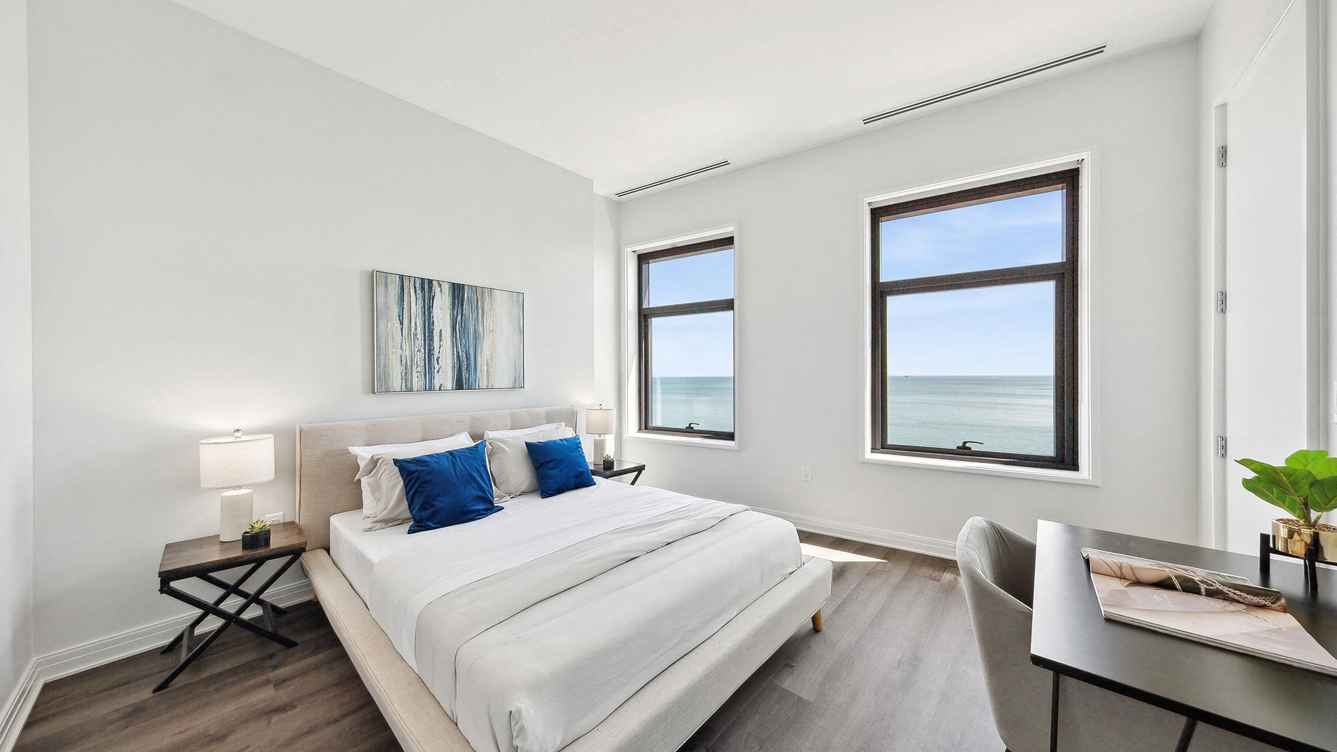 A minimalistic bedroom with a large bed, two windows with an ocean view, a desk, a chair, two bedside tables, and a painting above the bed.