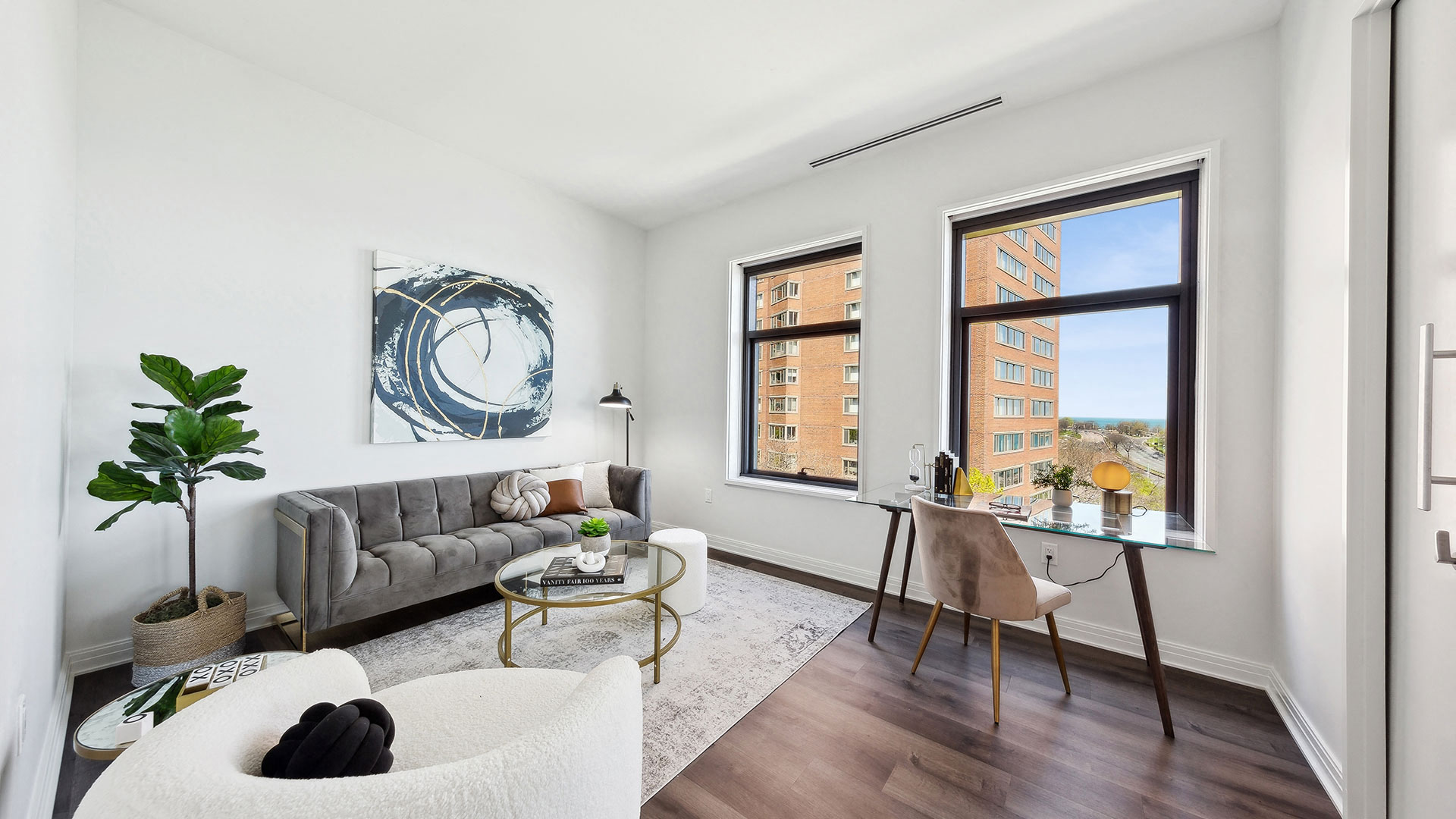 A modern living area with a gray sofa, white chairs, abstract wall art, a large plant, a small round coffee table, a desk with a chair by the window, and a view of a nearby building from two windows.