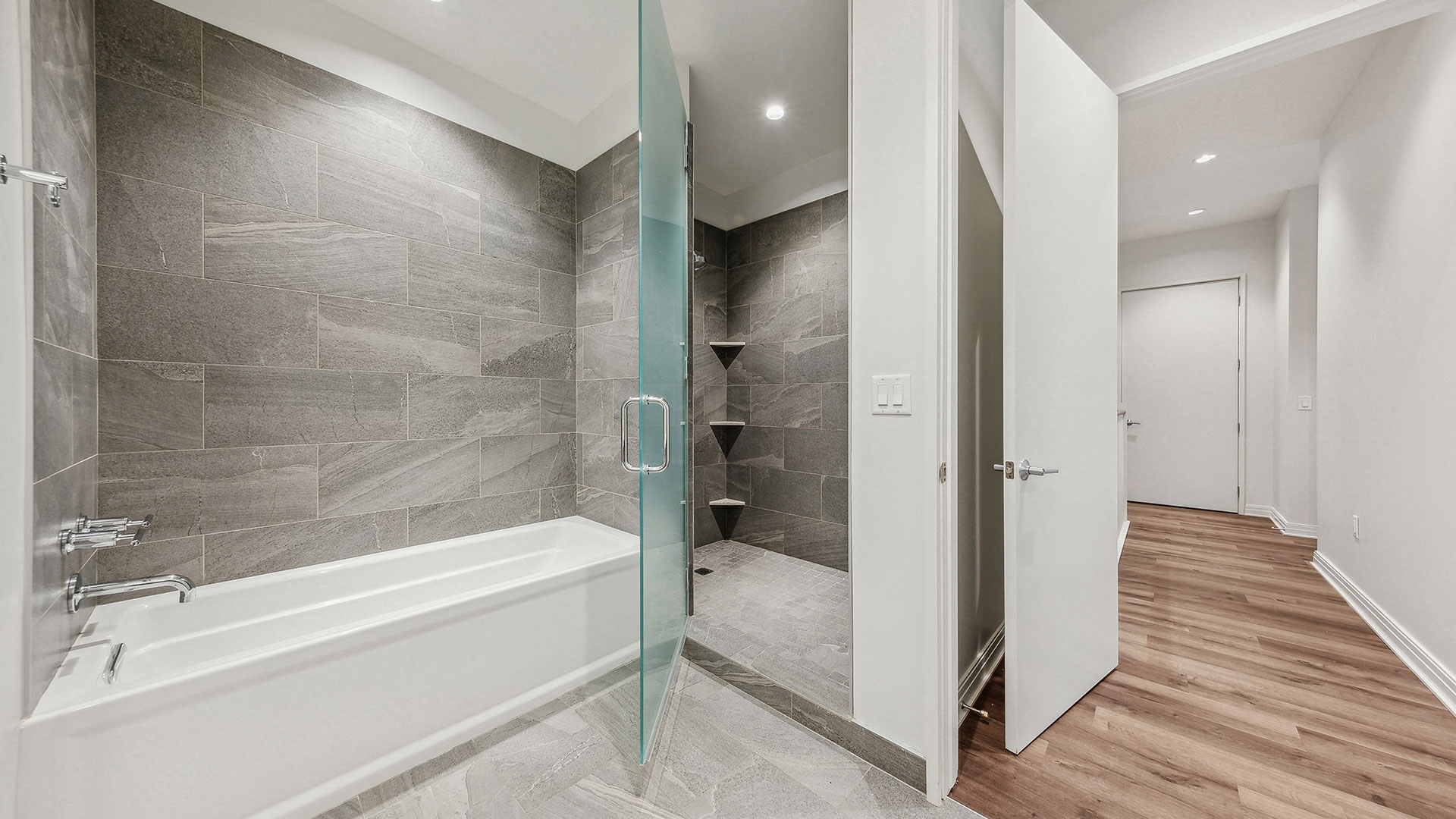 Modern bathroom with a bathtub, glass shower door, and grey-tiled walls. Hallway with wood flooring visible through an open door.