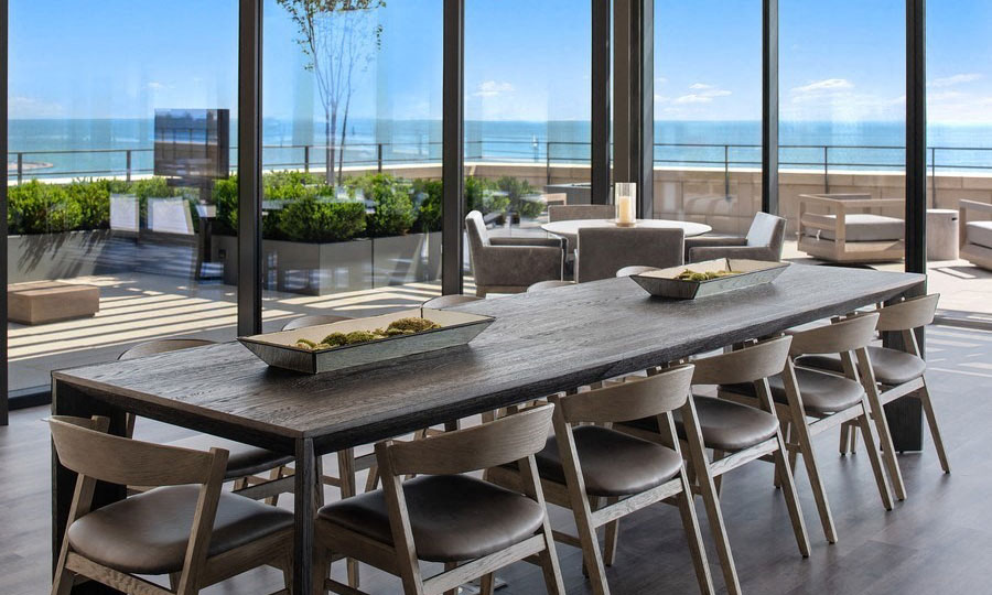 A long dinging room table sits in a open room with floor-to-celing windows. The sky terrace is seen outside and Lake Michigan beyond on a sunny day.