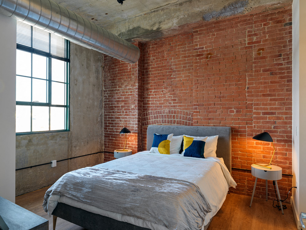 A bedroom with a brick wall and a bed with a grey comforter.