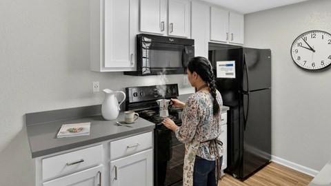 A woman is cooking in a kitchen with a black refrigerator and a clock on the wall.