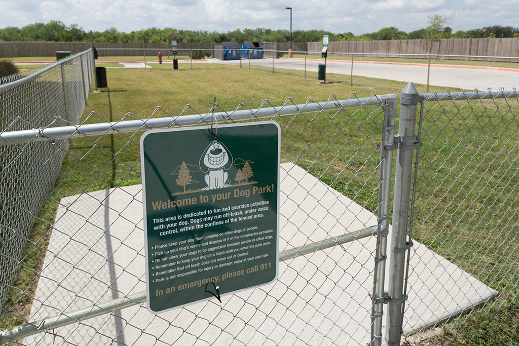 a sign hanging on a chain link fence with a dog park behind it