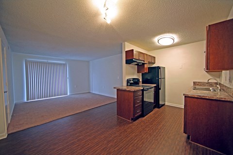 an empty kitchen and living room with wood flooring and a black refrigerator