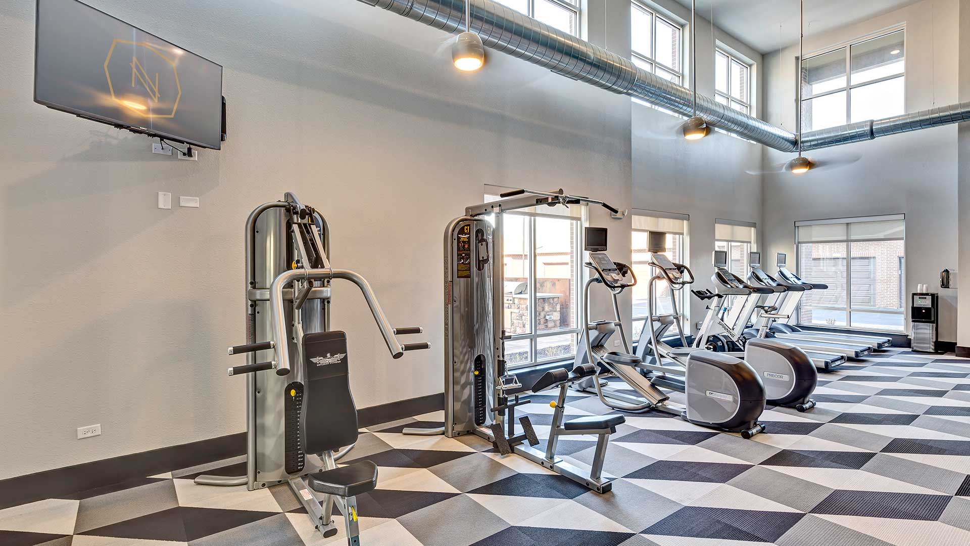 A row of fitness equipment is in front of windows.