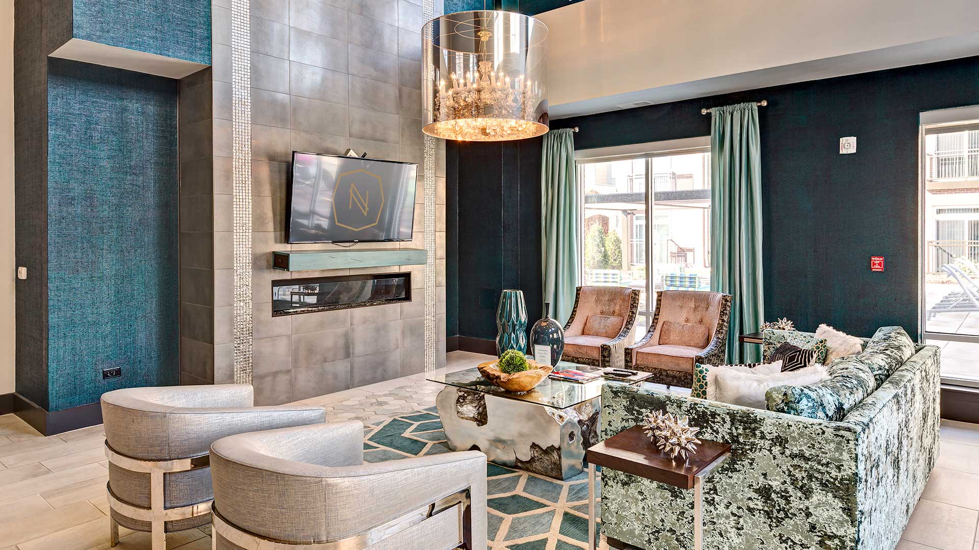 Various club furniture sits around a stone and glass coffee table facing a large television on the wall. A chandelier hangs above.