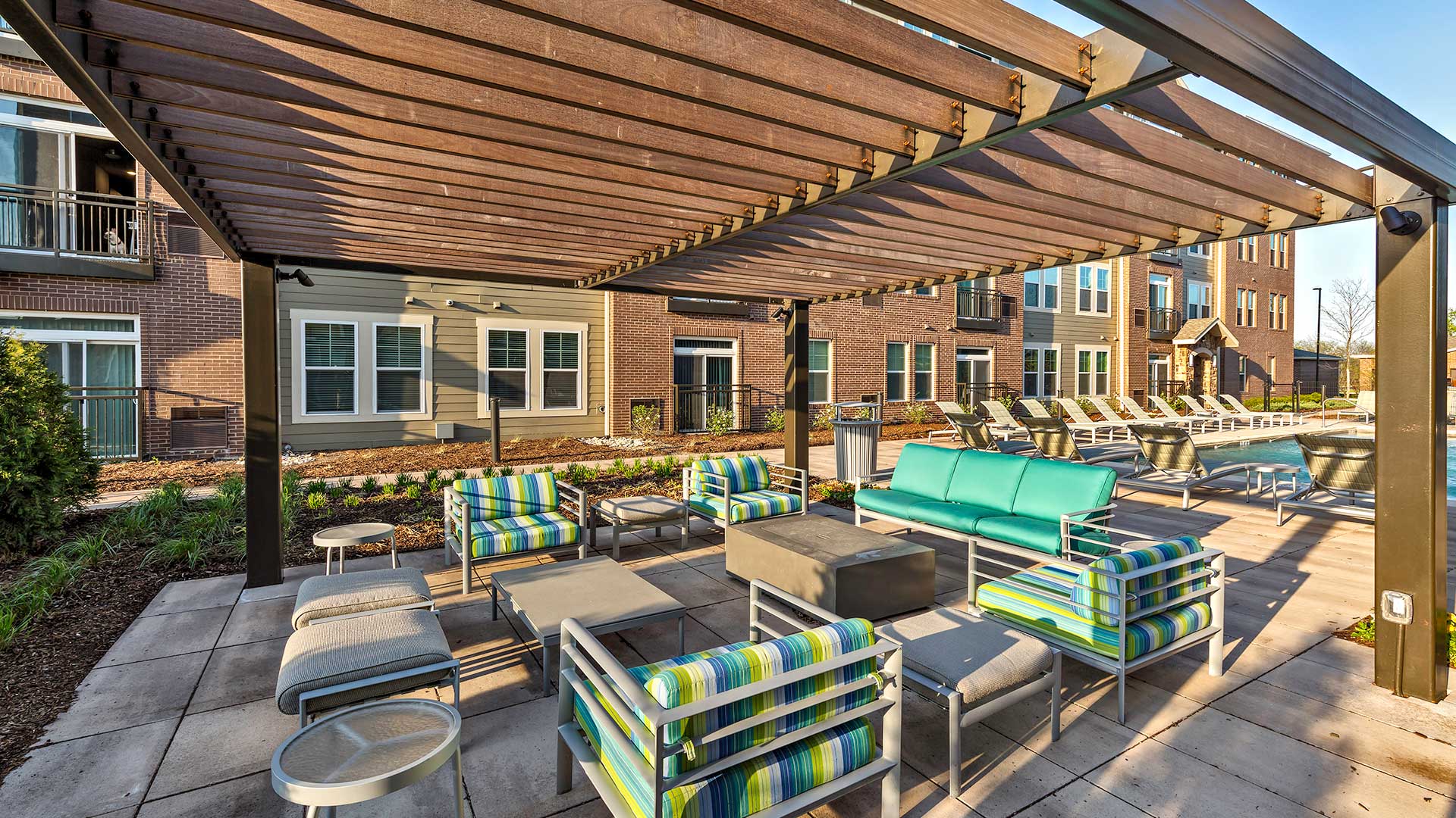 A large pergola with various outdoor seating around a rectangular table and a rectangular fire pit. The pool is seen behind to the right.