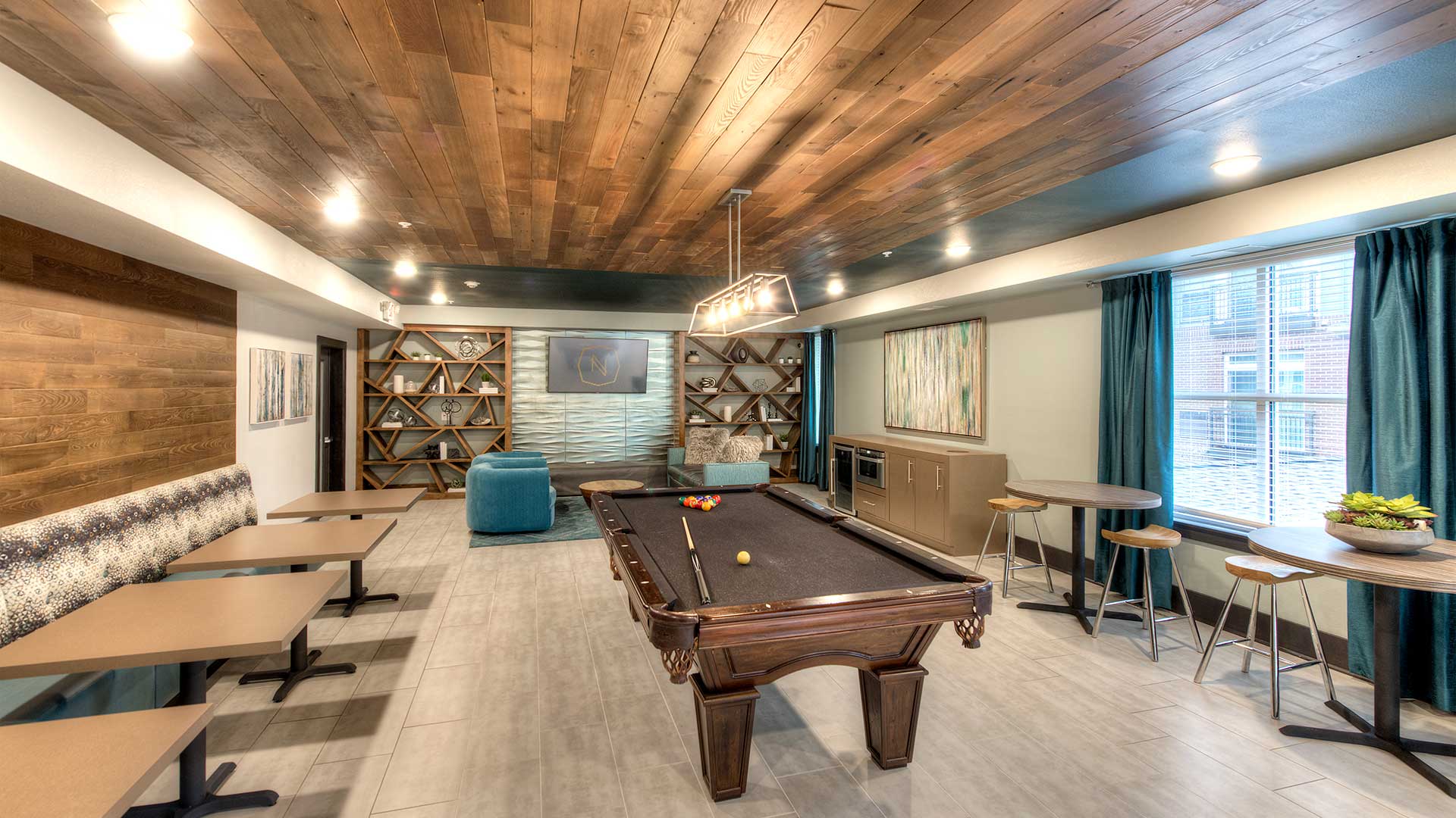 There is a pool table in the middle of the room. The left wall has bench seating with tables. The back wall is a seating area with couches. Café tables and a coffee bar line the right wall.