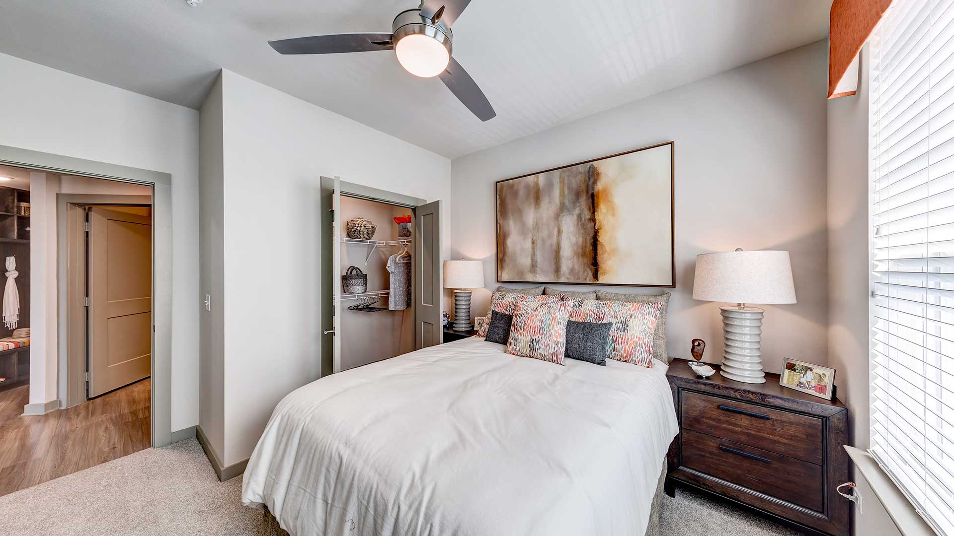 A large bed with an end table and lamp on either side. An open closet is on the left wall. On the far left is the doorway to the rest of the apartment.