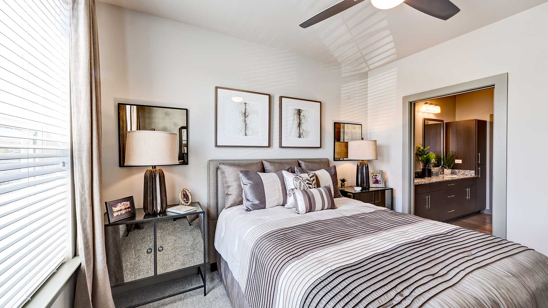 A large bed is set against the wall with an end table and lamp on each side. To the right is an opening that leads to the bathroom.