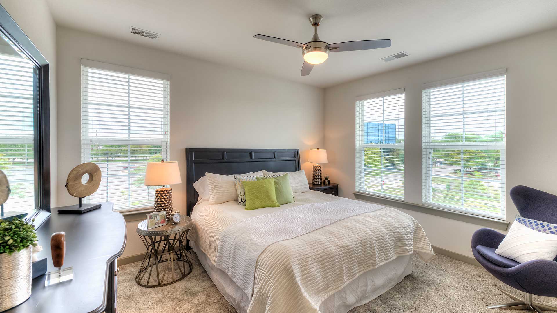 A bed in a carpeted room. There is a dresser on the left and chair on the right. The windows show a bright day outside.