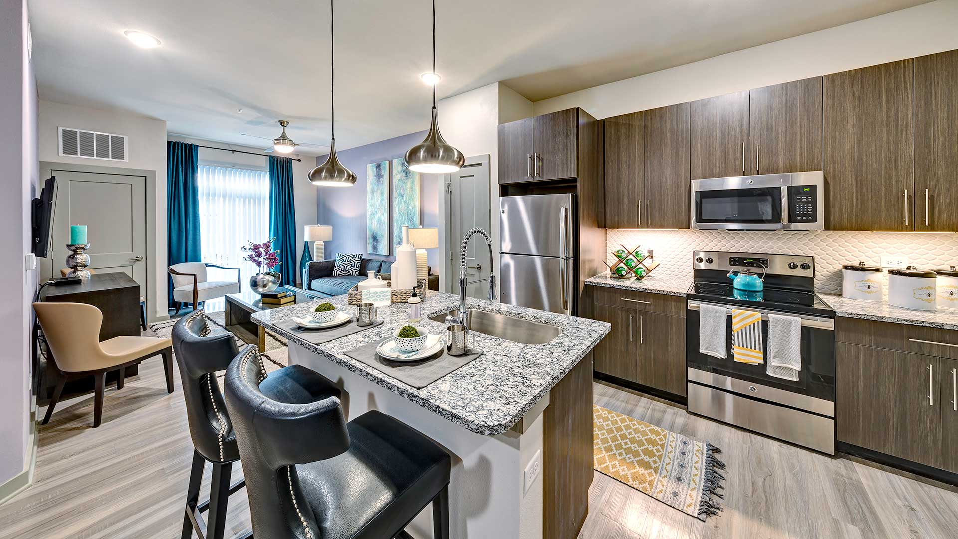 A kitchen in a residence at North680. The stainless steel appliances and brown cabinets are along the far wall. An island is in front with a built-in sink and two place settings. The living room is se