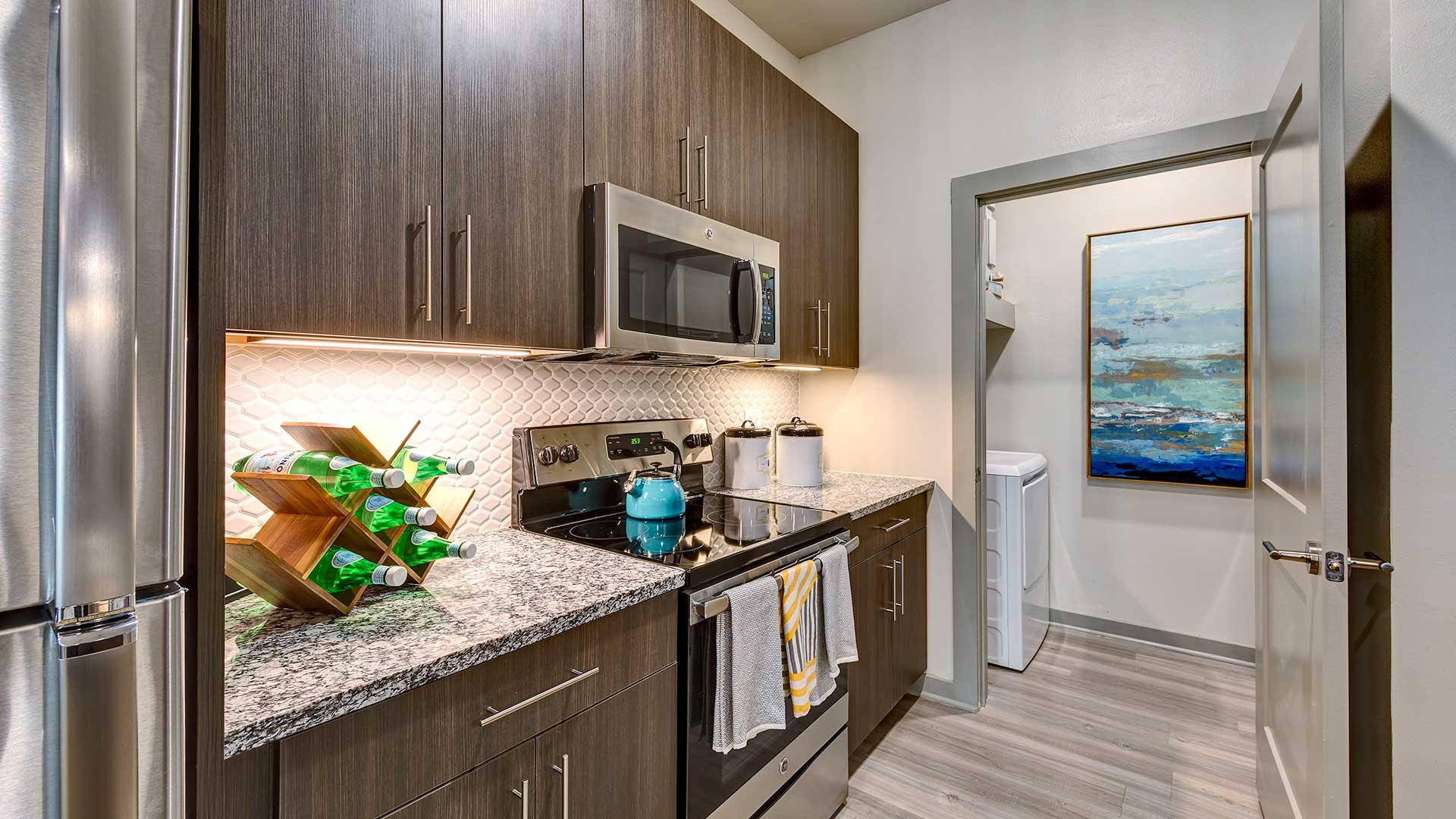 Looking through a kitchen with stainless steel appliances and brown cabinets into a walk-in closet with a dryer in view.