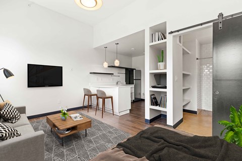 A living room with a grey couch, a coffee table, a television, and a kitchen area in the background.