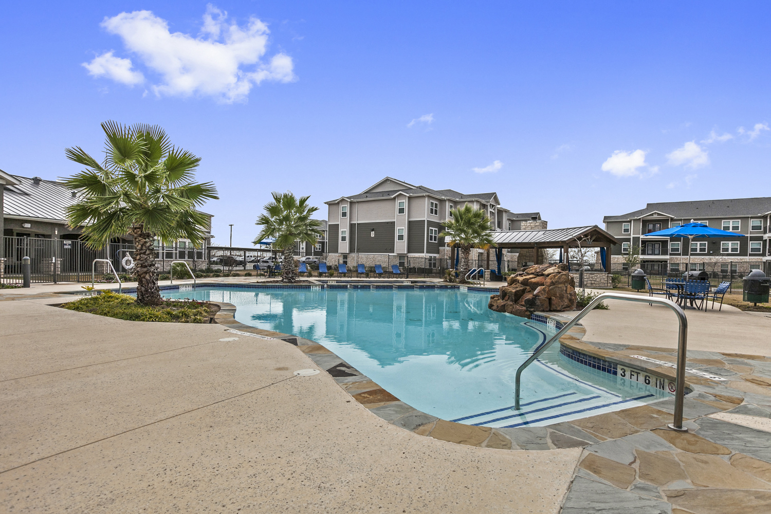 Apartments for Rent New Braunfels TX - Canyon House Apts - Swimming Pool with Rock Feature Surrounded by Concrete, Tile, and Two Small Palm Trees. Features