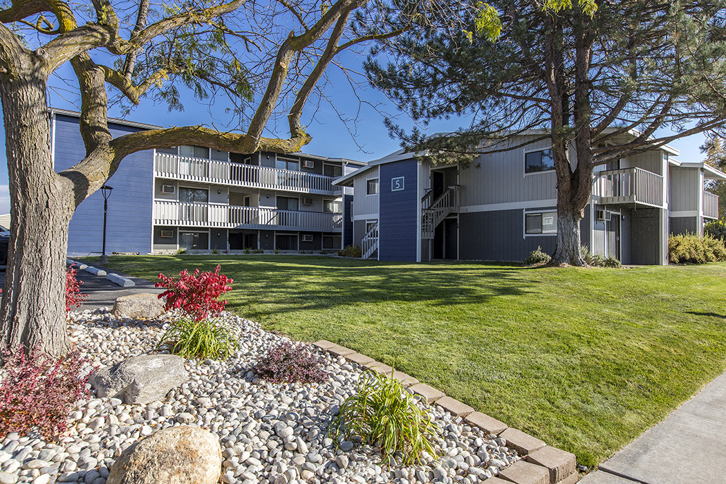 Exterior view of manicured grounds and building. Rock garden, large trees and blue skies in the background at The Lakes Apartments, Moses Lake, WA.