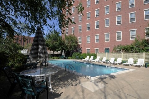 A pool surrounded by chairs and umbrellas in front of a red brick building.