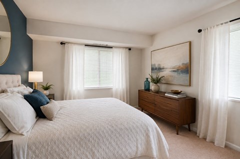A bedroom with a large bed, a nightstand with a lamp, a dresser with a plant, and a painting on the wall.