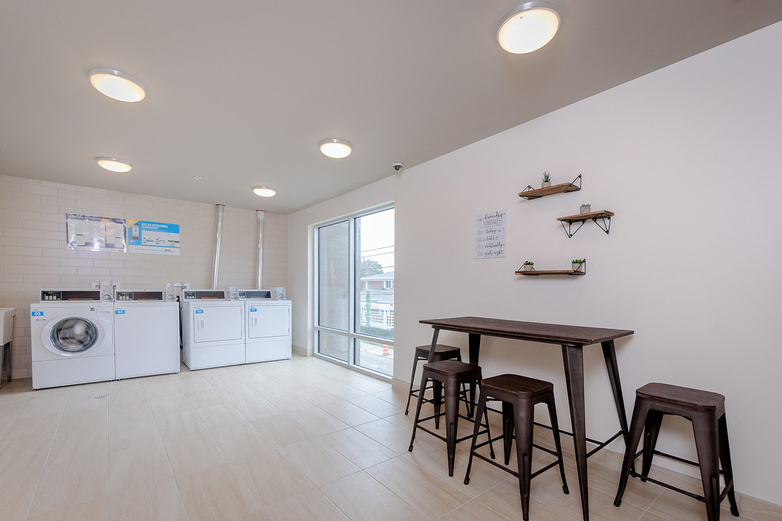 Laundry room area of an apartment, fitted with washing machines, seating area, and vinyl flooring.